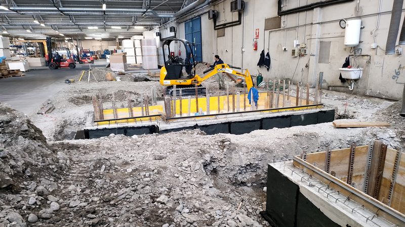 Interno di capannone industriale con scavo, casseforme e armature per il getto di nuove fondazioni, con miniescavatore al lavoro.